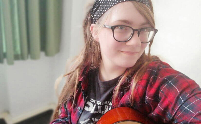 junge Frau mit Brille und Haarband und Gitarre in der Hand